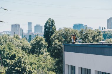 arka planda yeşil ağaçlar ve mavi gökyüzü ile çatı üzerinde dururken yükseltilmiş elleriyle gesturing afrikalı amerikalı adam 