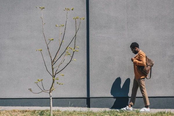 灰色の壁に沿って日当たりの良い通りを歩くスタイリッシュなアフリカ系アメリカ人ビジネスマンの側面図 — ストック写真
