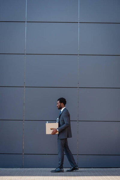 side view of fired african american businessman carrying carton box while walking along wall