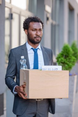 dismissed african american businessman looking away while holding carton box clipart