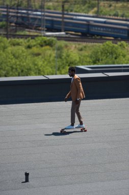 arka planda demiryolları ve trenler ile güneşli çatı üzerinde longboard sürme Afrikalı Amerikalı işadamı