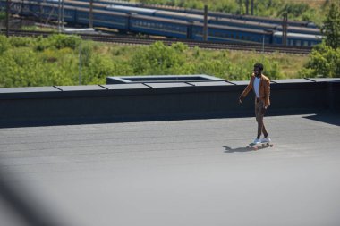 selective focus of african american businessman riding longboard on sunny rooftop with railways and trains on background clipart