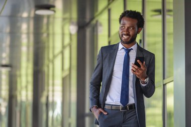 smiling african american businessman with smartphone walking near office building and holding hand in pocket clipart