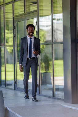 ofis binası yakınında yürüyen akıllı telefon tutan gülümseyen Afrikalı Amerikalı işadamı 