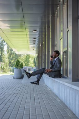 ofis binası yakınında otururken akıllı telefon konuşurken neşeli afrikalı-Amerikalı işadamı
