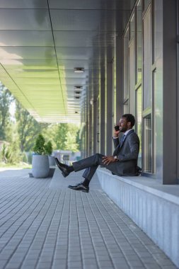 cheerful african american businessman talking on smartphone while sitting near office building clipart