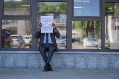 african american businessman reading economic news newspaper while sitting near office building with glass facade clipart