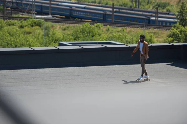 鉄道や電車を背景に日当たりの良い屋上にロングボードに乗るアフリカ系アメリカ人ビジネスマンの選択的な焦点 — ストック写真