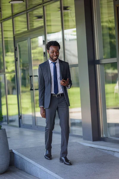 Uśmiechnięty African American Biznesmen Trzyma Smartphone Podczas Spaceru Wzdłuż Biura — Zdjęcie stockowe