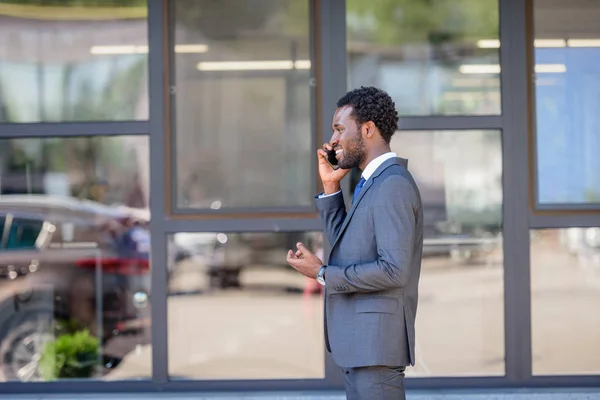 オフィスビルの近くに立ちながらスマートフォンで話す陽気なアフリカ系アメリカ人ビジネスマン — ストック写真