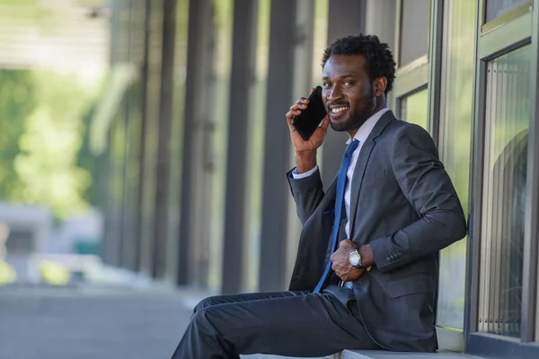 陽気なアトリカンアメリカのビジネスマンは スマートフォンで話し パラペットに座っている間 カメラで笑顔 — ストック写真