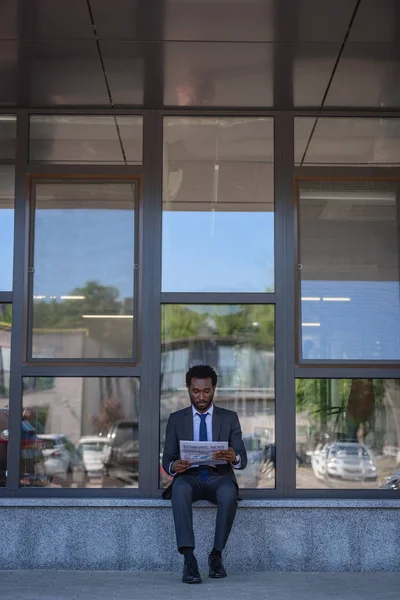 ガラスのファサードでオフィスビルの近くに座っている間 ハンサムなアフリカ系アメリカ人のビジネスマンが新聞を読んでいます — ストック写真