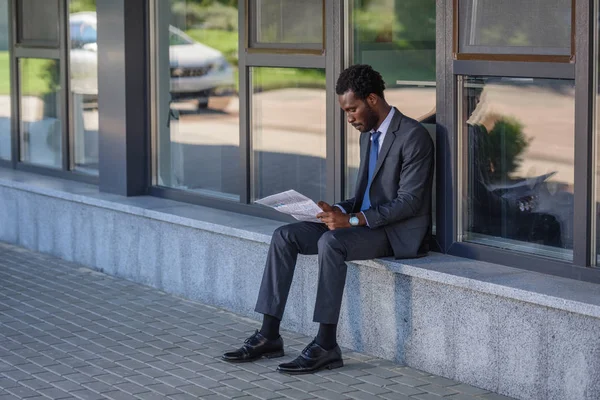 Skoncentrowany African American Biznesmen Czytania Gazety Siedząc Parapet Budynku Biurowego — Zdjęcie stockowe