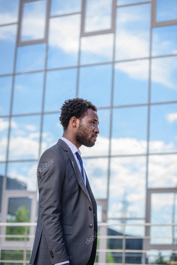 confident african american businessman in suit walking along building with glass facade