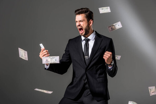 excited businessman with smartphone near falling dollar banknoteson on grey