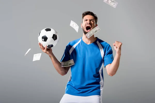 soccer player with ball cheering with clenched hand near falling money on grey