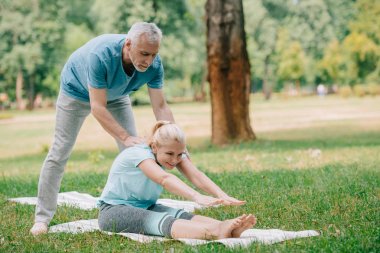 handsome mature man helping woman practicing yoga on lawn in park clipart