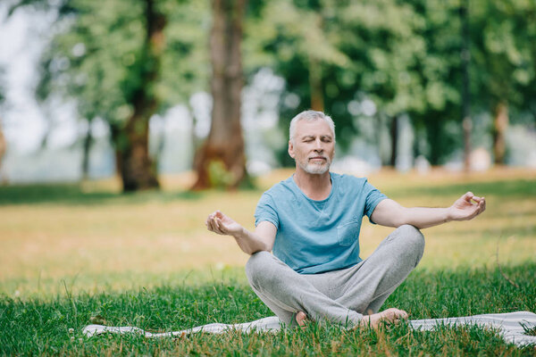 handsome mature man meditating while sitting in lotus pose in park