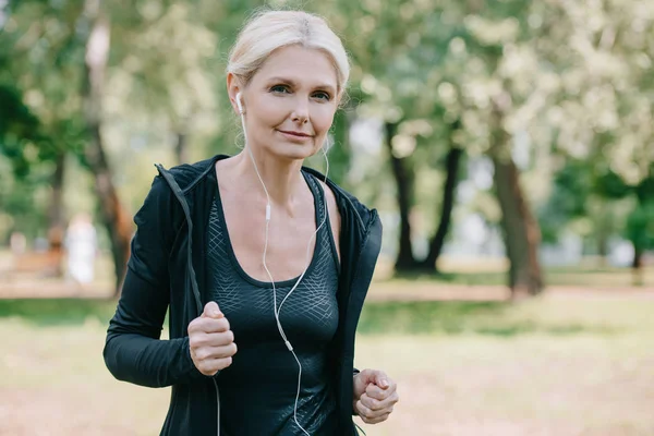 Hermosa Deportista Madura Sonriendo Cámara Mientras Corre Parque Escuchar Música — Foto de Stock