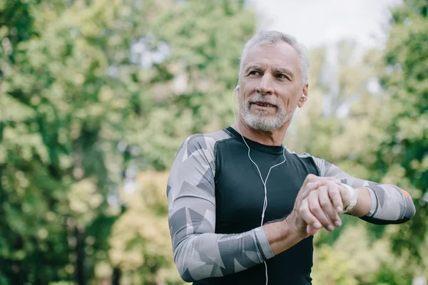 Smiling Mature Sportsman Touching Fitness Tracker While Listening Music Earphones — Stock Photo, Image