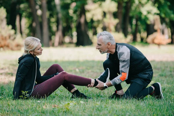 Attentive Mature Sportsman Touching Injured Leg Sportswoman Sitting Lawn — Stock Photo, Image