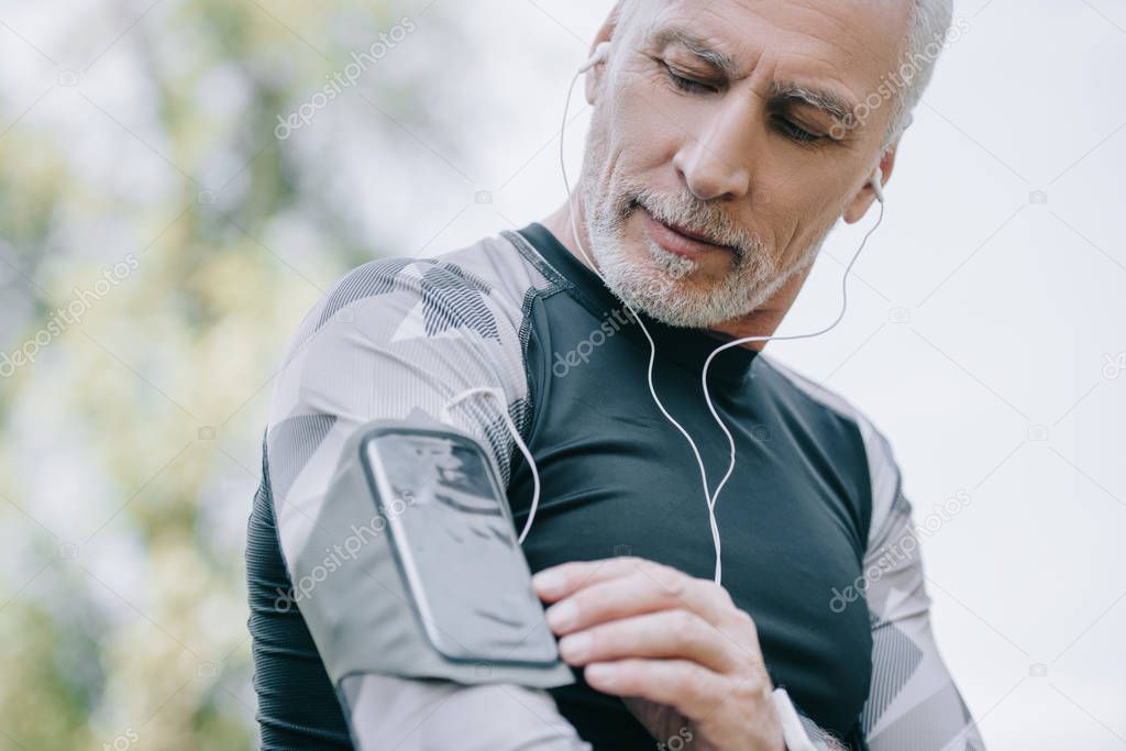 handsome mature sportsman using smartphone in armband case and listening music in earphones