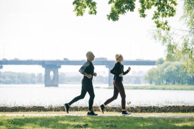 side view of sportive mature joggers running along riverside in park clipart