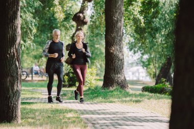 selective focus of smiling mature sportsman and sportswoman running together in park clipart