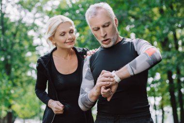 surprised mature sportsman looking at fitness tracker near smiling sportswoman clipart
