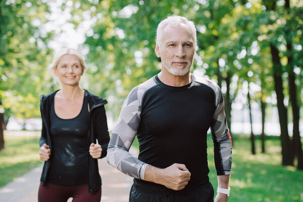 mature, smiling sportsman and sportswoman running in park together 