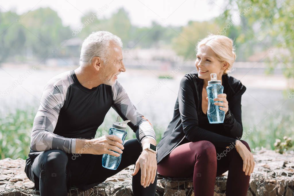 cheerful mature sportsman and sportswoman looking at each other while sitting on parapet with sports bottles