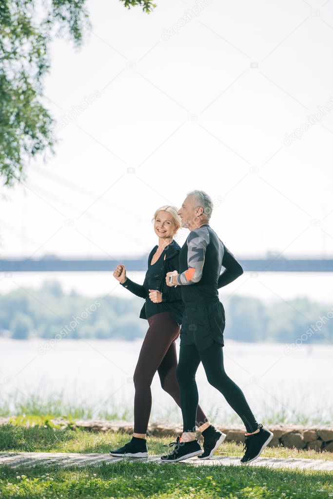 cheerful mature sportswoman running along riverside in park near handsome sportsman