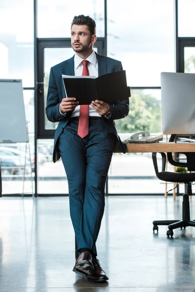 Guapo Hombre Negocios Sosteniendo Carpeta Mientras Está Pie Oficina Moderna —  Fotos de Stock