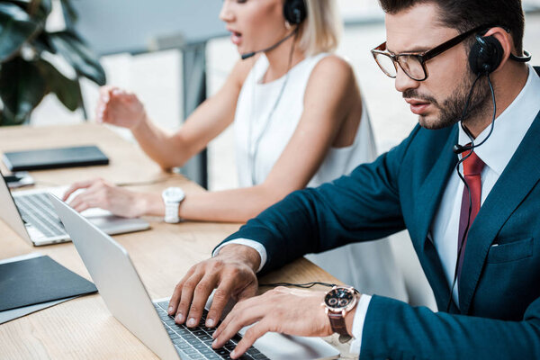 selective focus of handsome man in glasses typing on laptop near coworker in headset 