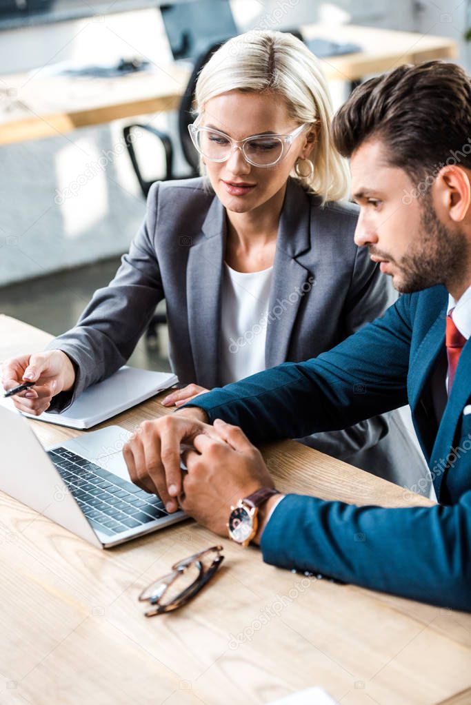 attractive woman in glasses holding pen near laptop near bearded man in office 