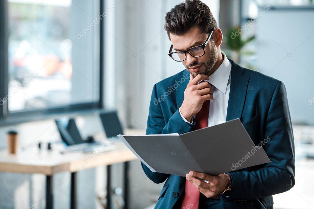 pensive businessman in glasses looking at folder and touching face 