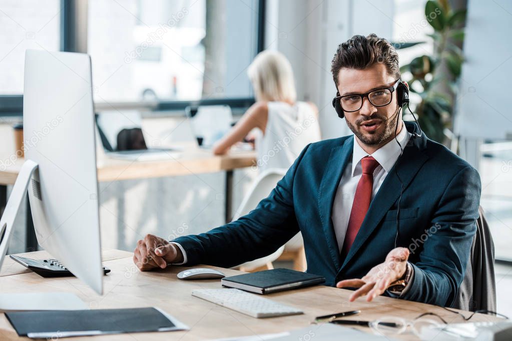 selective focus of bearded businessman in glasses and headset gesturing in office 