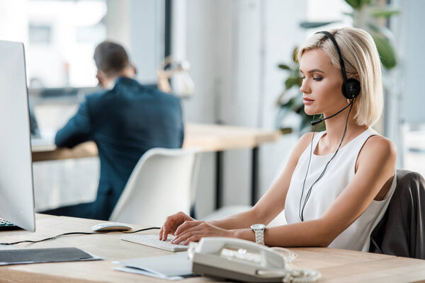 selective focus of attractive blonde businessman in headset typing on computer keyboard in office 