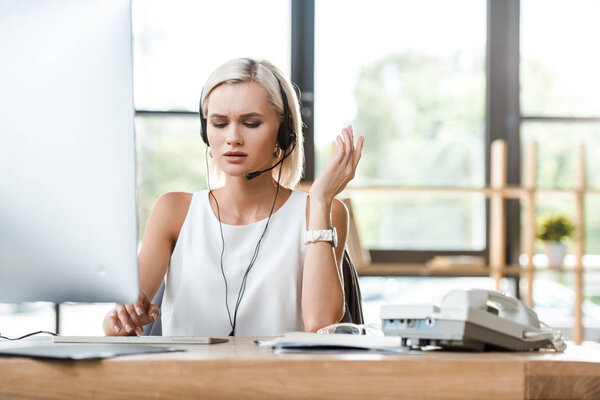 selective focus of upset blonde woman in headset gesturing while working in office 