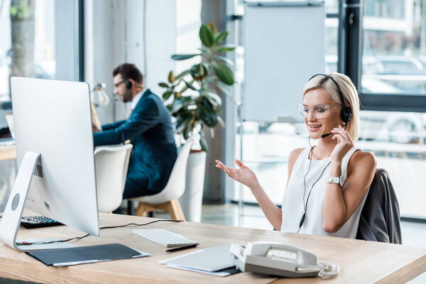 selective focus of cheerful blonde woman in headset gesturing while talking near coworker 