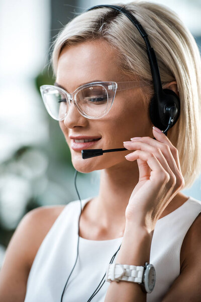 close up of cheerful blonde operator in glasses touching headset 