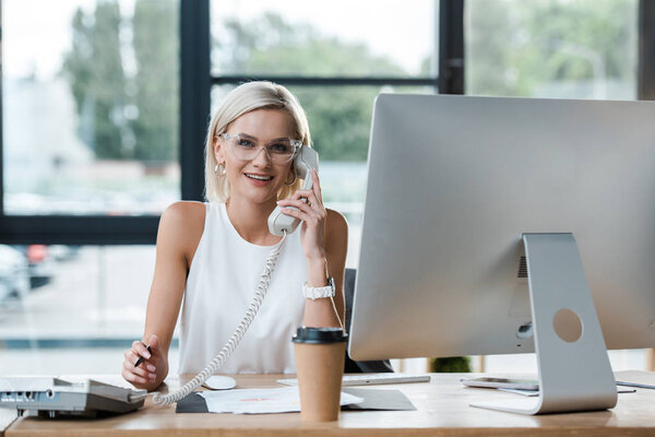 selective focus of happy blonde businesswoman talking on retro phone in office 
