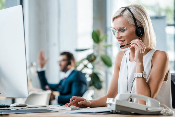 selective focus of happy operator typing on computer keyboard near coworker 