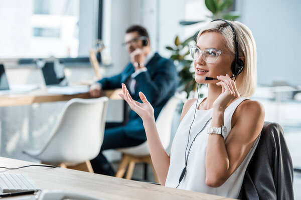 selective focus of happy operator touching headset, talking and gesturing near coworker 