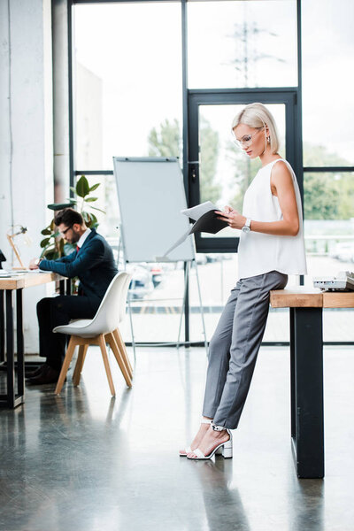 selective focus of attractive businesswoman in glasses holding folder and documents near coworker in modern office 
