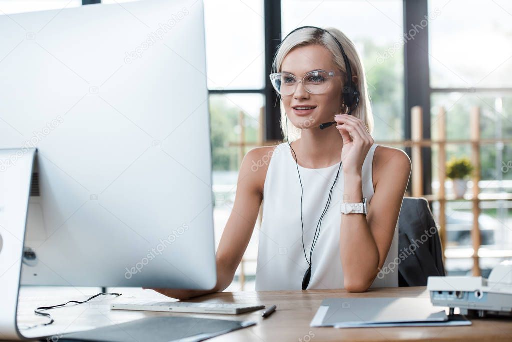 selective focus of cheerful blonde woman in headset working in office near computer monitor 