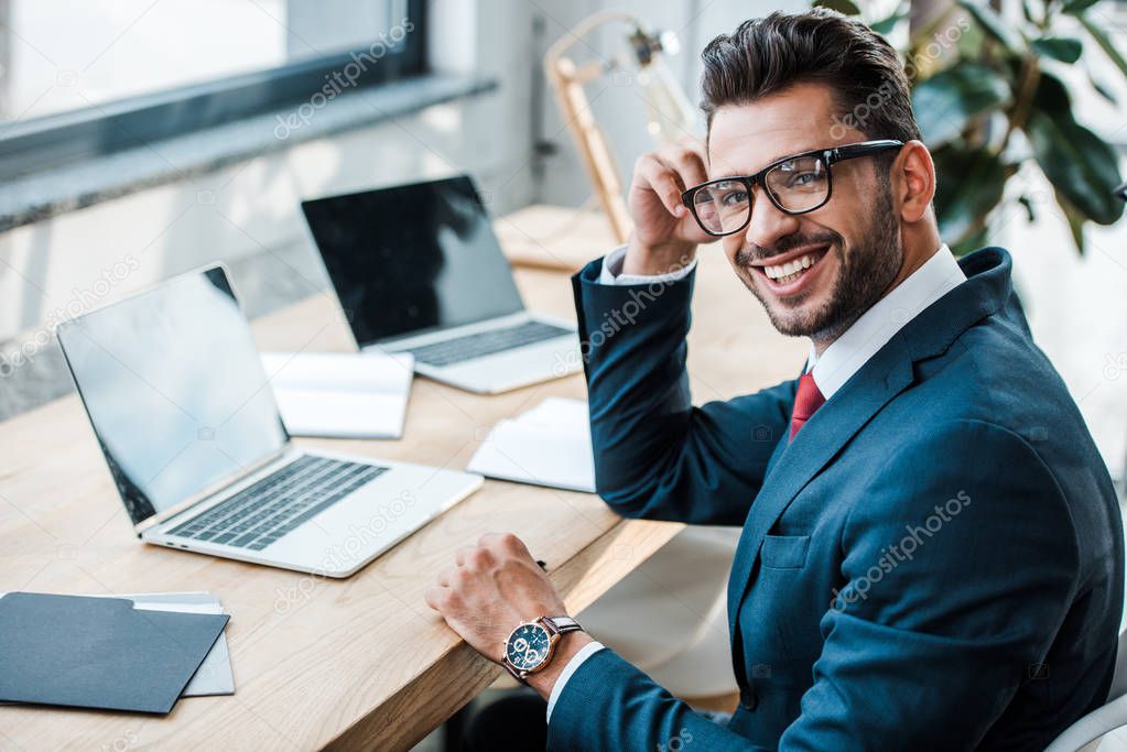 selective focus of cheerful man in glasses looking at camera near laptops with blank screens 