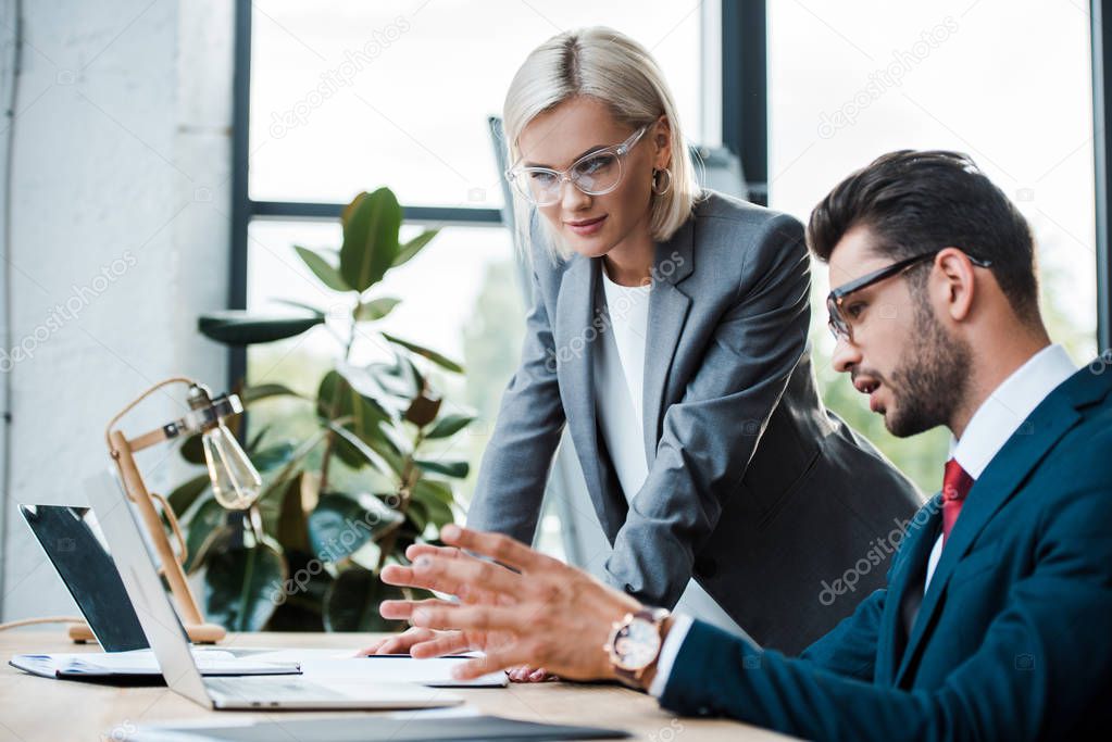 selective focus of bearded man gesturing near laptop and blonde woman in glasses 