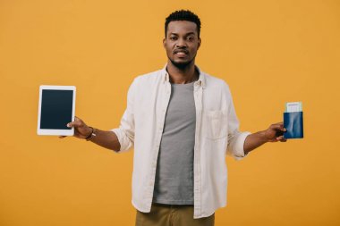 turuncu izole boş ekran ve pasaport ile dijital tablet tutan duygusal afro-amerikan adam