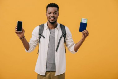 turuncu izole boş ekran ile uçak bileti ve akıllı telefon ile pasaport tutan sırt çantası ile mutlu afrikalı amerikalı adam